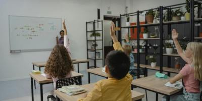 Diverse group of students raising hands in a vibrant classroom setting with teacher at front.